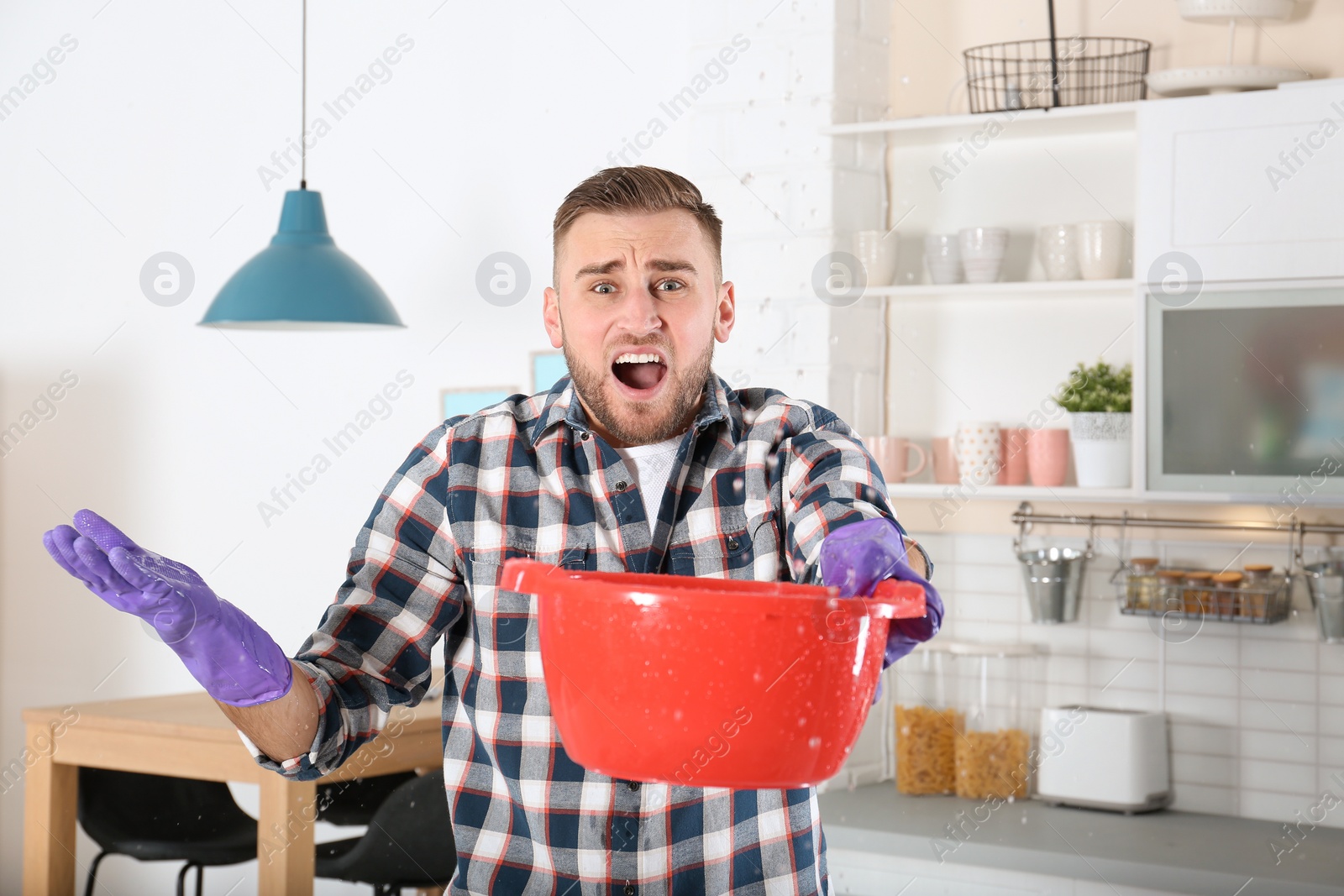 Photo of Emotional young man holding plastic basin under water leakage from ceiling in kitchen. Plumber service