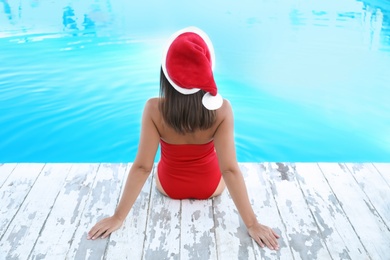 Photo of Young woman wearing Santa Claus hat near swimming pool, back view. Christmas vacation