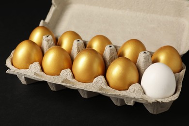 Photo of Ordinary chicken egg among golden ones in box on black background, closeup