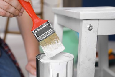 Photo of Man dipping brush into can of white paint indoors, closeup