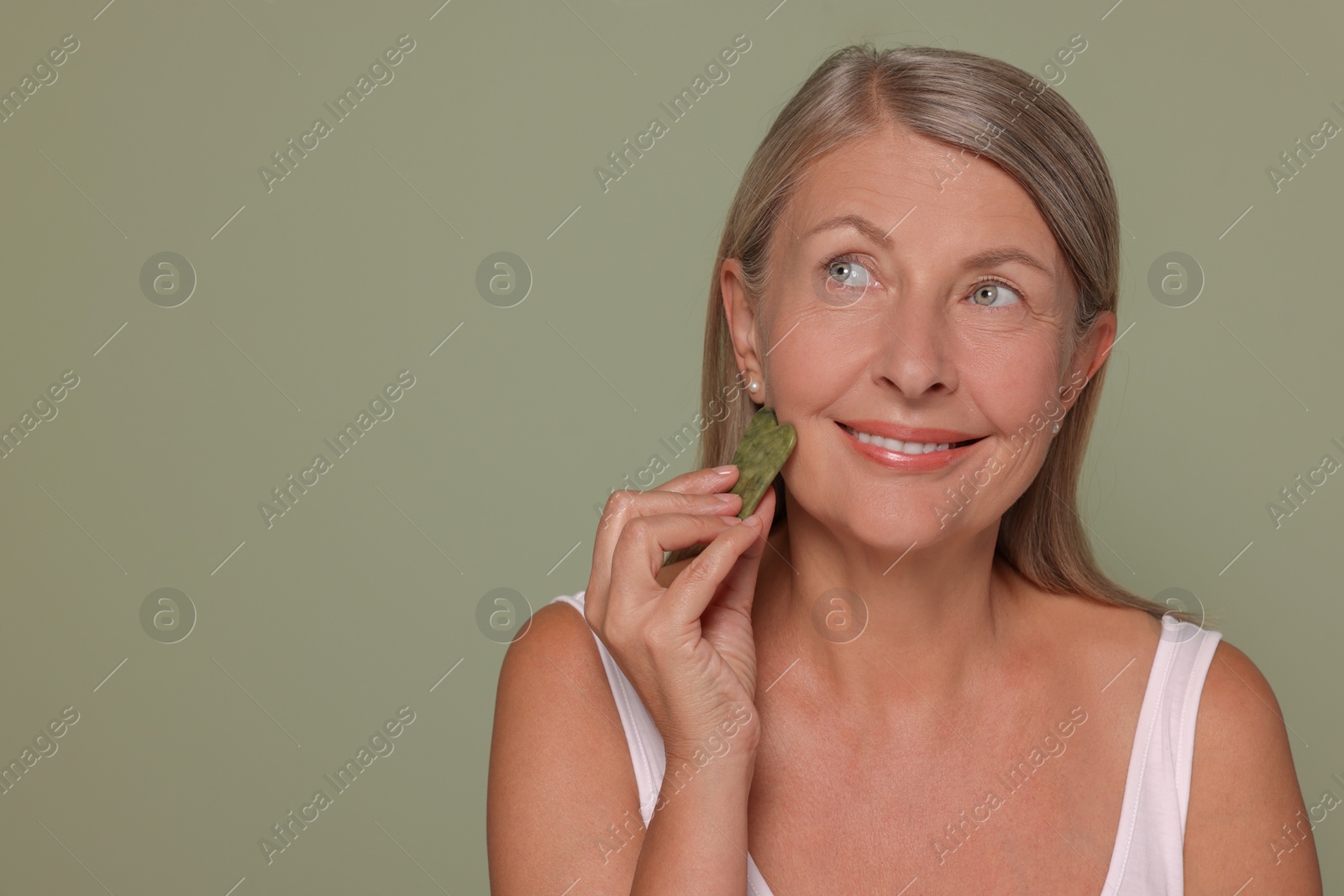 Photo of Woman massaging her face with jade gua sha tool on green background. Space for text