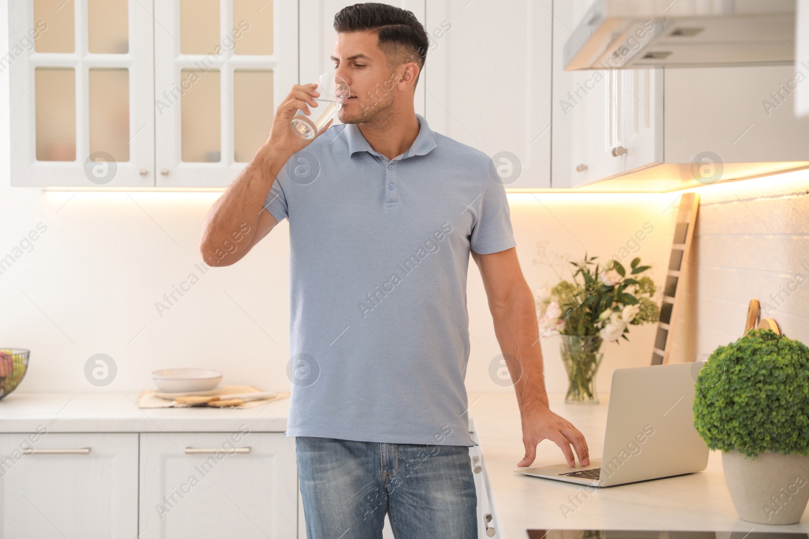 Photo of Man drinking glass of pure water while working on laptop in kitchen