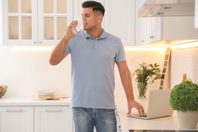 Man drinking glass of pure water while working on laptop in kitchen