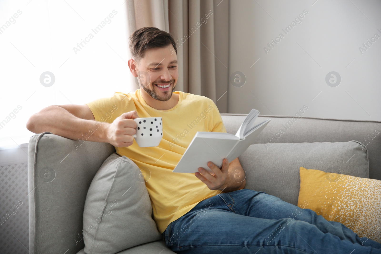 Photo of Handsome man with cup of coffee reading book on sofa at home