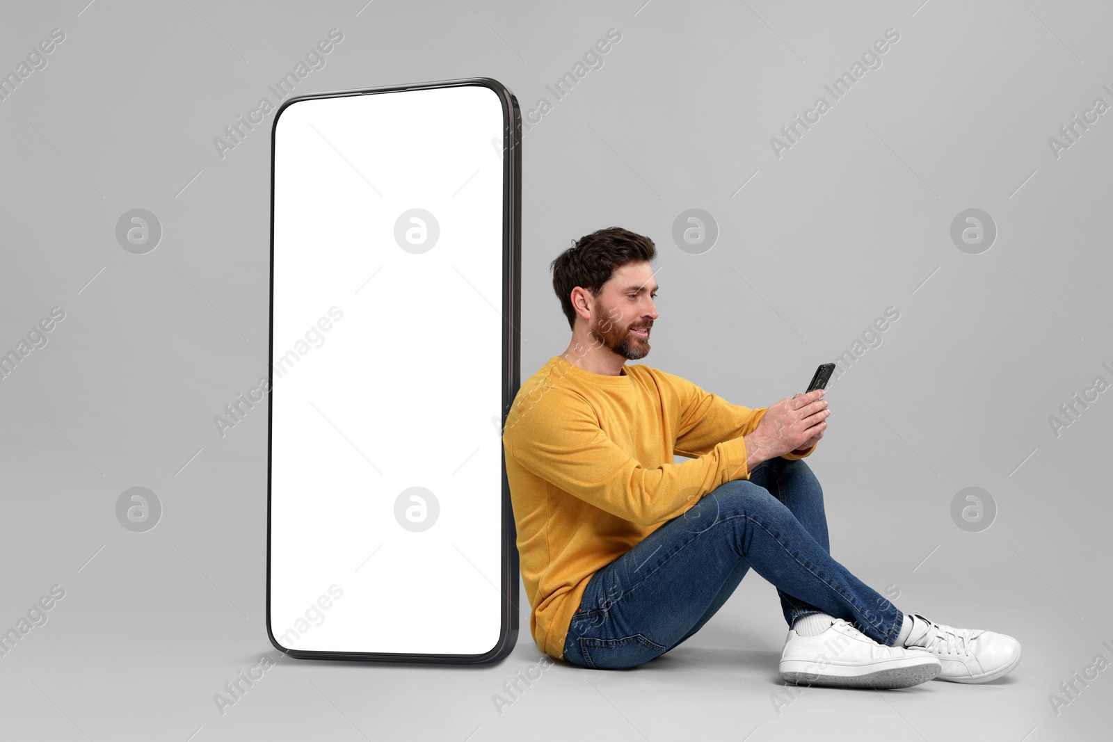 Image of Man with mobile phone sitting near huge device with empty screen on grey background. Mockup for design