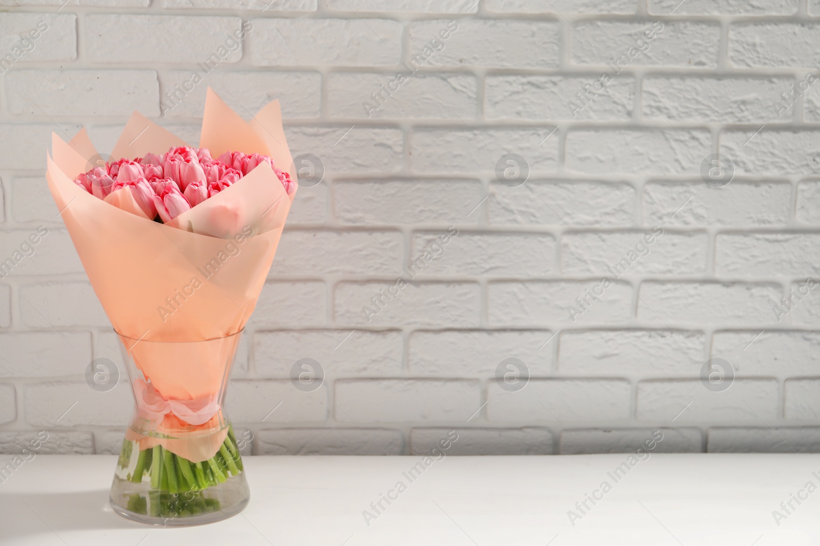 Photo of Bouquet of beautiful pink tulips in vase on white table near brick wall, space for text