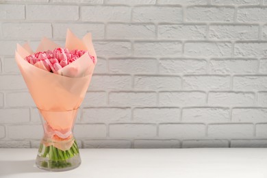 Bouquet of beautiful pink tulips in vase on white table near brick wall, space for text