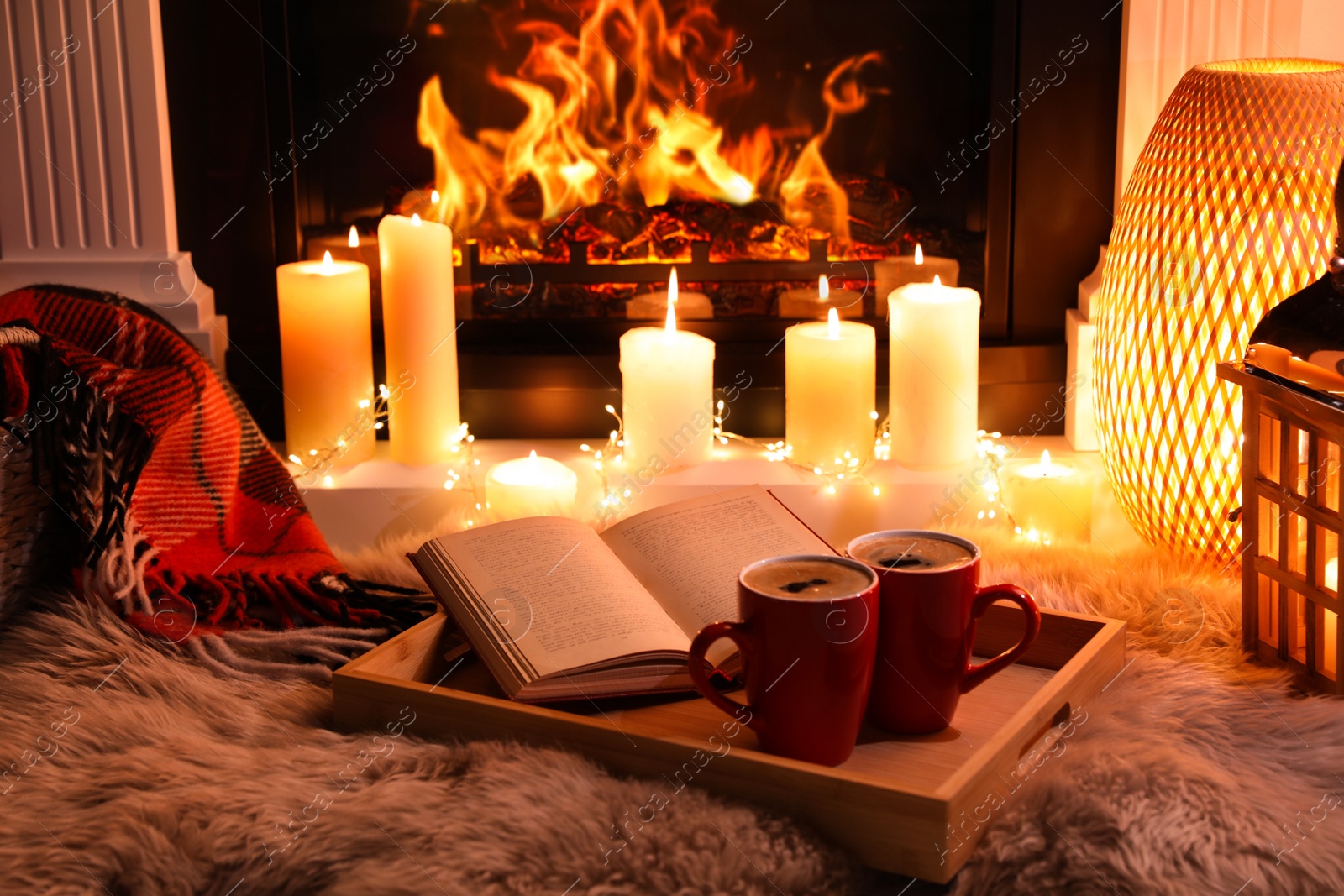 Photo of Cups of hot drink and open book near fireplace at home