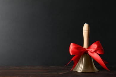 Photo of Golden bell with red bow on wooden table near blackboard, space for text. School days