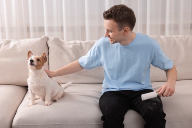 Photo of Pet shedding. Smiling man with lint roller removing dog's hair from pants on sofa at home
