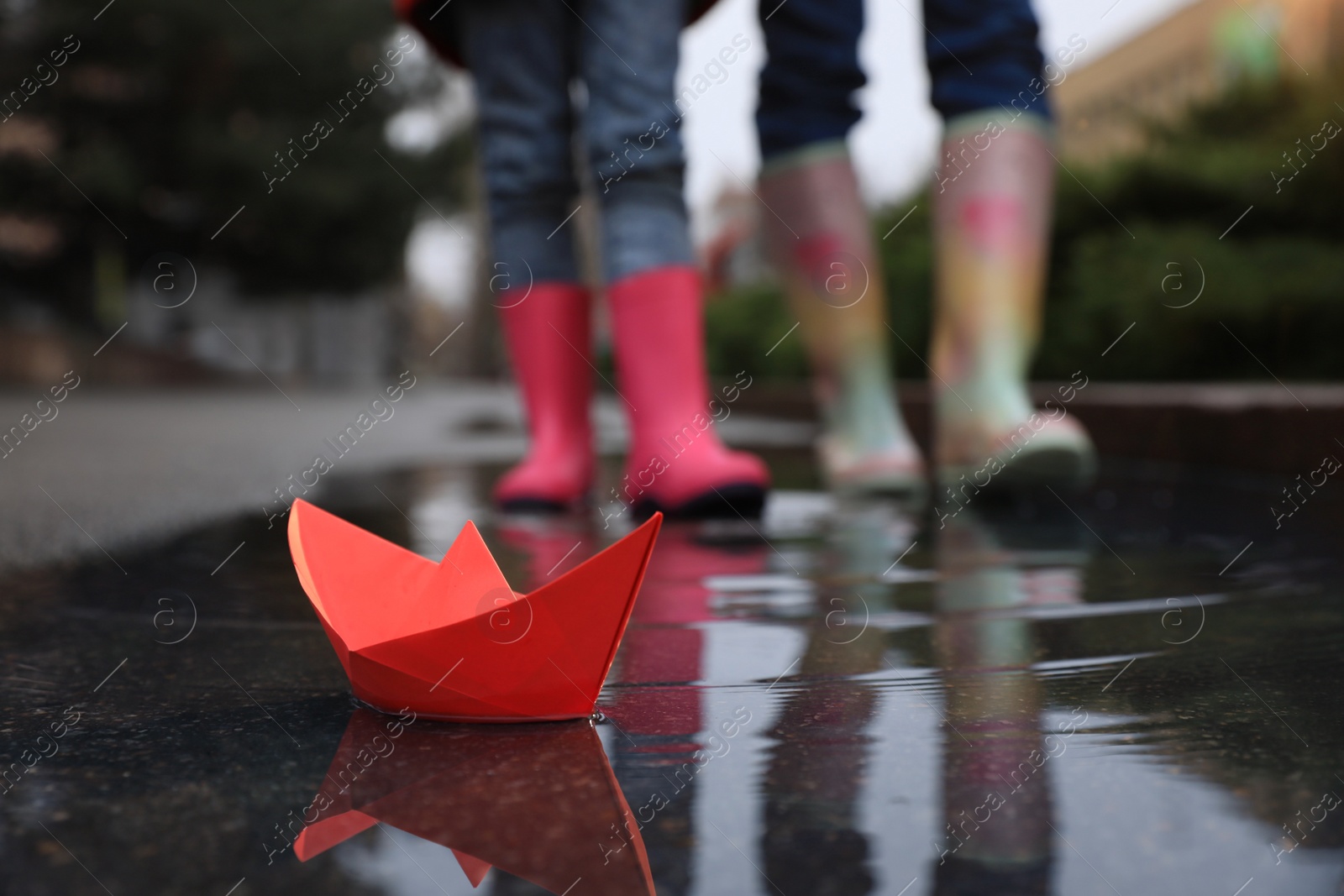 Photo of Little girl with her mother walking outdoors, focus on  paper boat in puddle