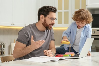 Photo of Father working remotely while his son playing with toys on desk at home. Man having video chat via laptop