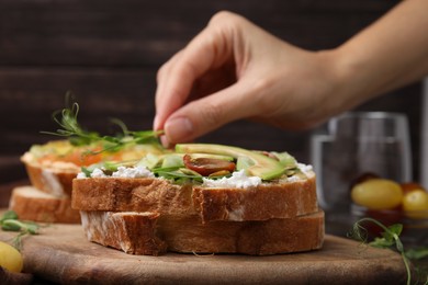 Photo of Woman putting microgreens on tasty vegan sandwich with avocado and tomato at table, closeup