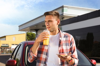 Photo of Young man with doughnut drinking juice near car at gas station