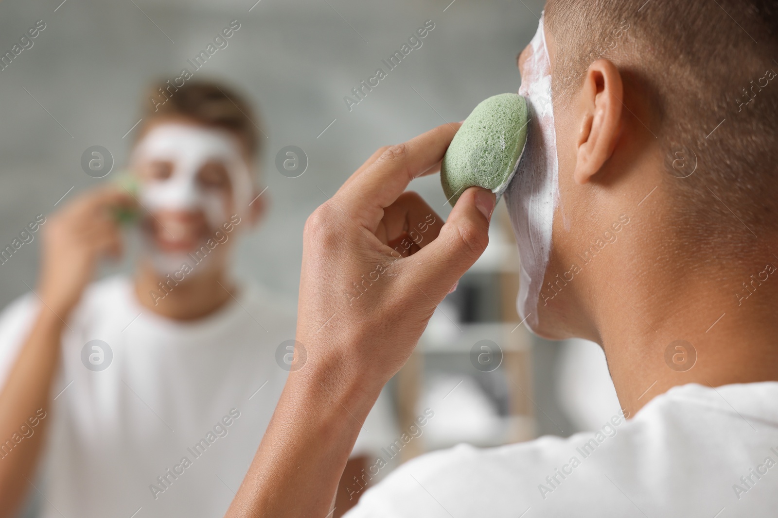 Photo of Young man washing off face mask with sponge near mirror in bathroom, closeup. Space for text