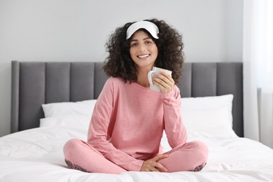 Photo of Beautiful young woman in stylish pyjama and sleep mask with cup of drink on bed at home