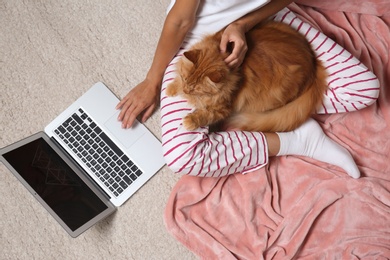 Photo of Woman with cute red cat and laptop on light carpet, top view