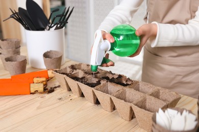 Photo of Little girl spraying water onto vegetable seeds in peat pots at wooden table indoors, closeup