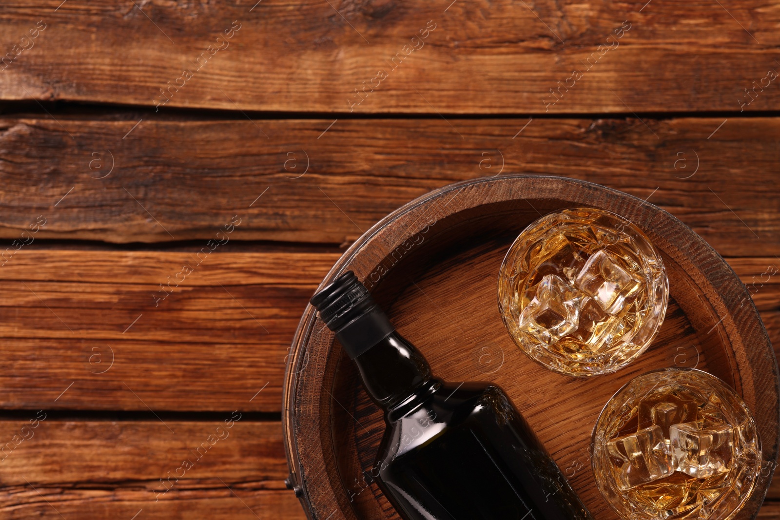 Photo of Whiskey with ice cubes in glasses, bottle and barrel on wooden table, top view. Space for text