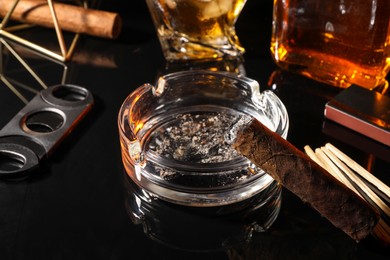Photo of Burnt cigar, ashtray, cutter and whiskey on black mirror surface, closeup