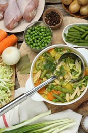 Saucepan of delicious vegetable soup with chicken and different ingredients on wooden table, flat lay