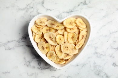 Photo of Heart shaped plate with banana slices on marble table, top view. Dried fruit as healthy snack