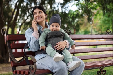Photo of Mother holding her child in sling (baby carrier) while talking on smartphone outdoors. Space for text