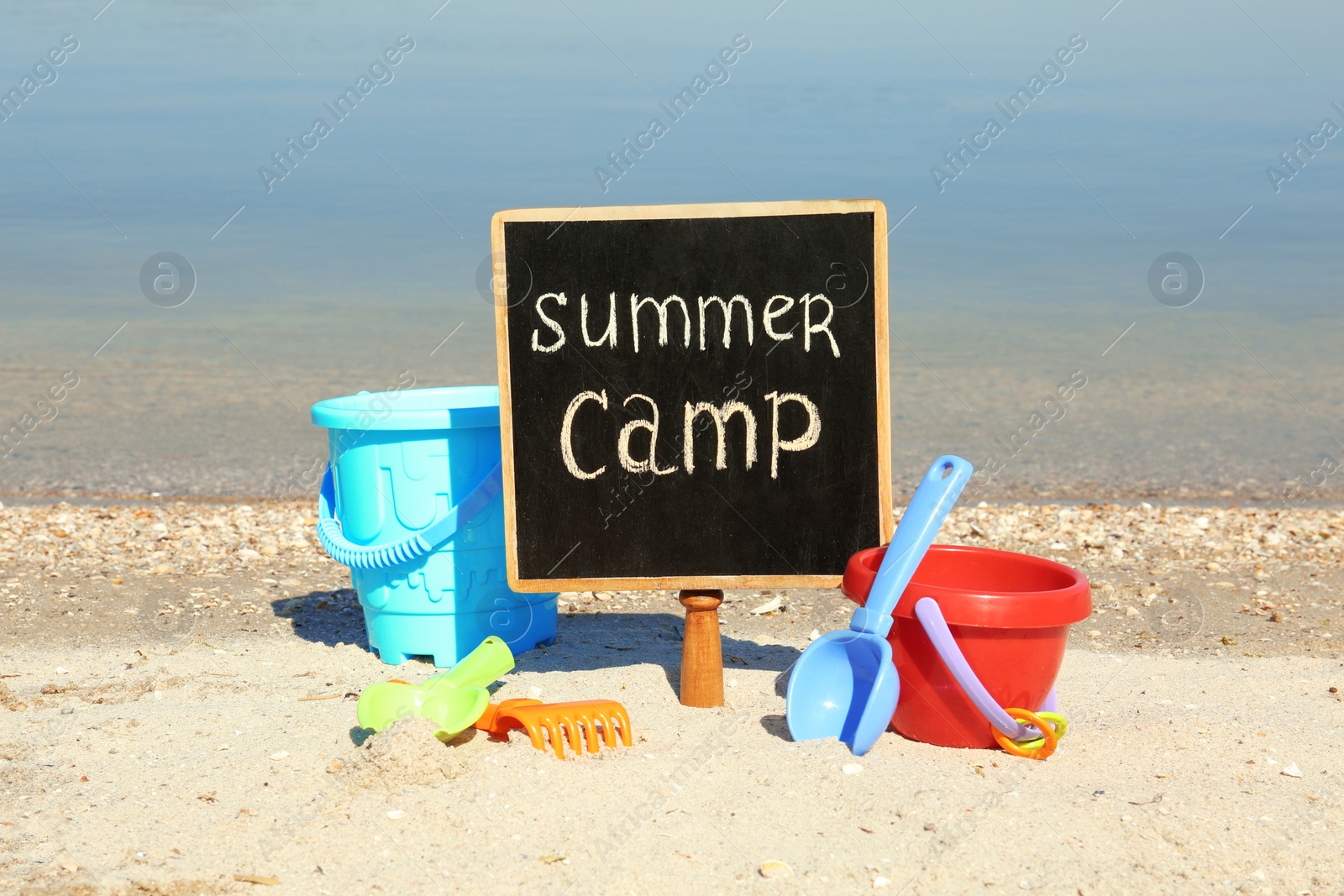 Photo of Small chalkboard with text SUMMER CAMP and beach toys on sand near river