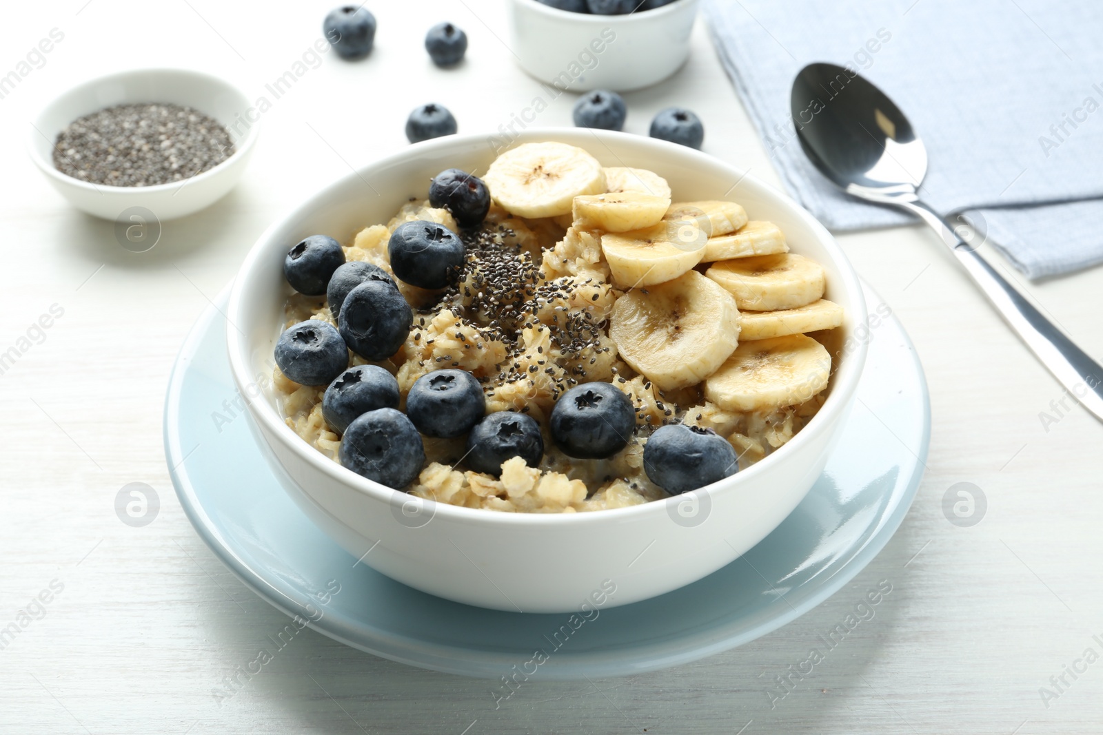 Photo of Tasty oatmeal with banana, blueberries and chia seeds served in bowl on white wooden table