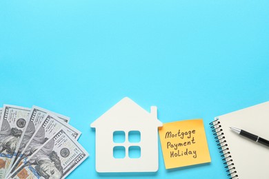 Photo of Note with words Mortgage Payment Holiday, house model, notebook and money on light blue background, flat lay. Space for text
