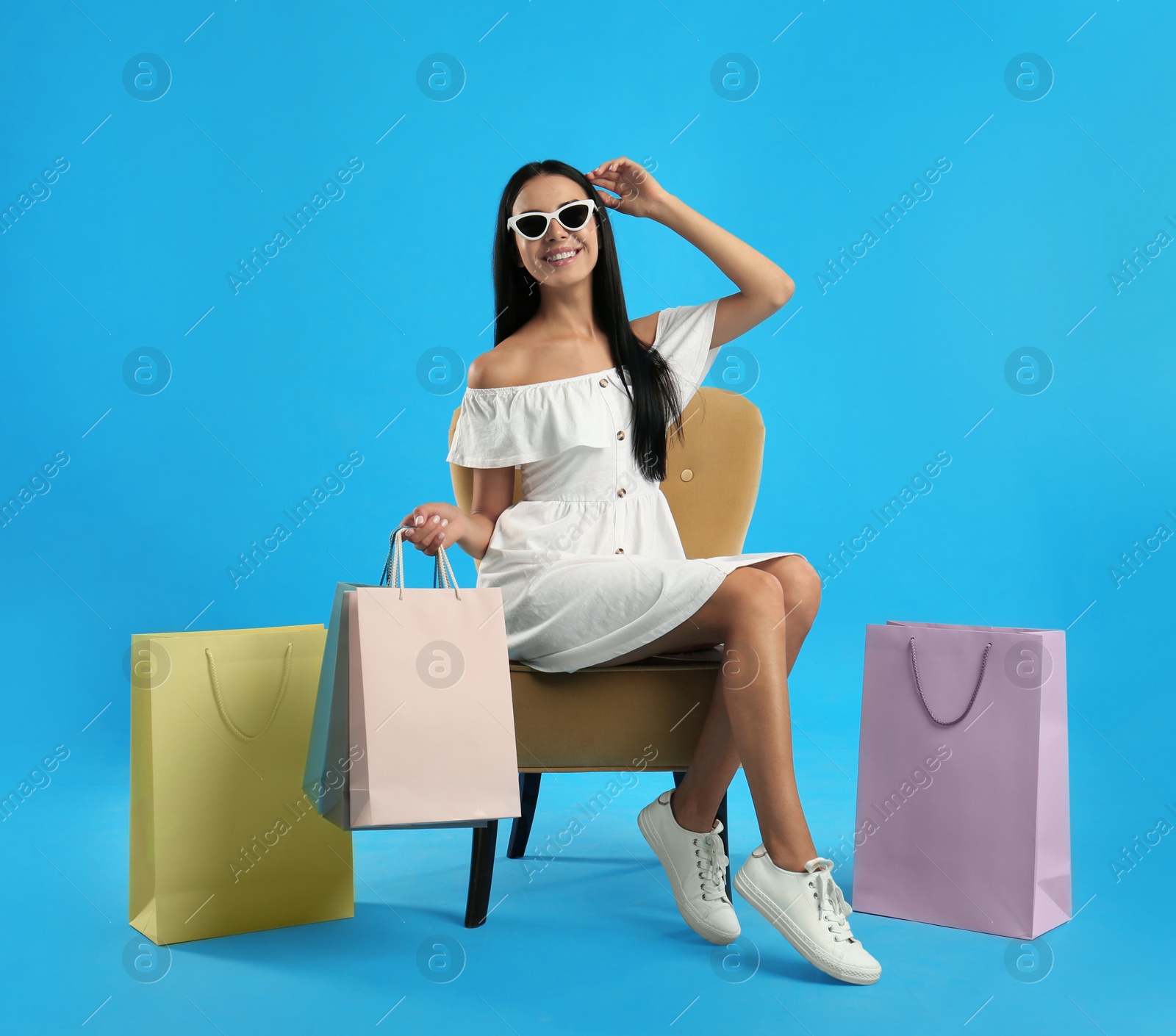 Photo of Beautiful young woman with paper shopping bags in armchair on light blue background