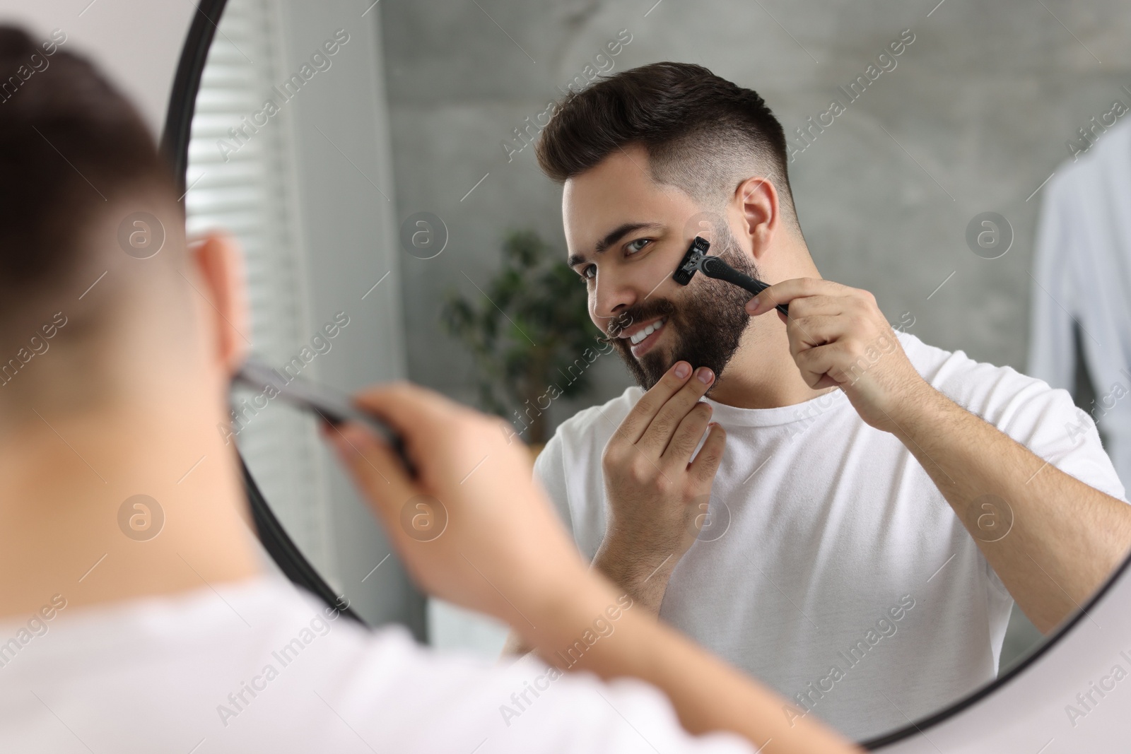 Photo of Handsome young man shaving with razor near mirror in bathroom