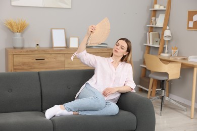 Woman waving hand fan to cool herself on sofa at home