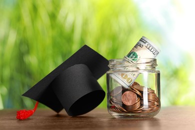 Photo of Scholarship concept. Glass jar with coins, dollar banknotes and graduation cap on wooden table