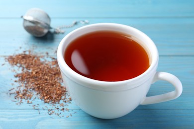 Photo of Ceramic cup of aromatic rooibos tea, infuser and scattered dry leaves on light blue wooden table, closeup