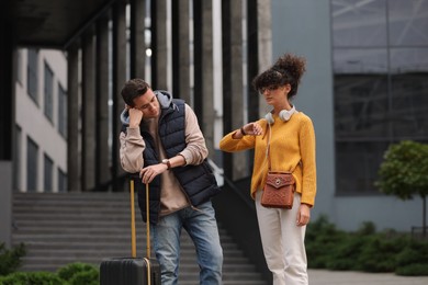Photo of Being late. Worried woman looking at watch and man with suitcase near building outdoors