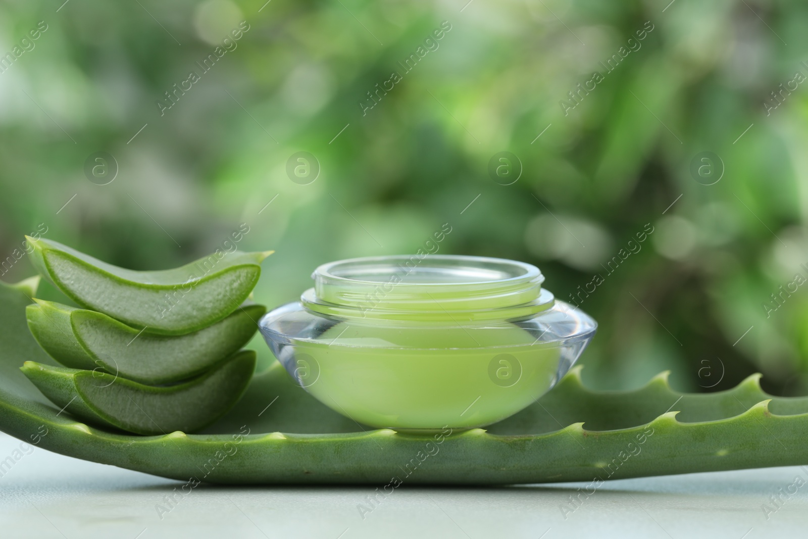 Photo of Jar with cream and cut aloe leaf on white table against blurred green background, closeup. Space for text