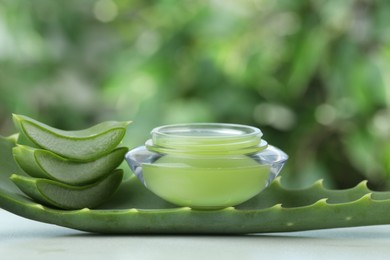 Jar with cream and cut aloe leaf on white table against blurred green background, closeup. Space for text