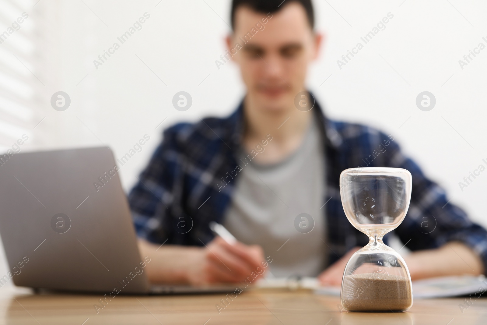 Photo of Hourglass with flowing sand on desk. Man taking notes while using laptop indoors, selective focus