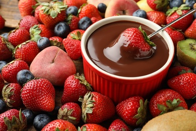 Fondue fork with strawberry in bowl of melted chocolate surrounded by other fruits on table