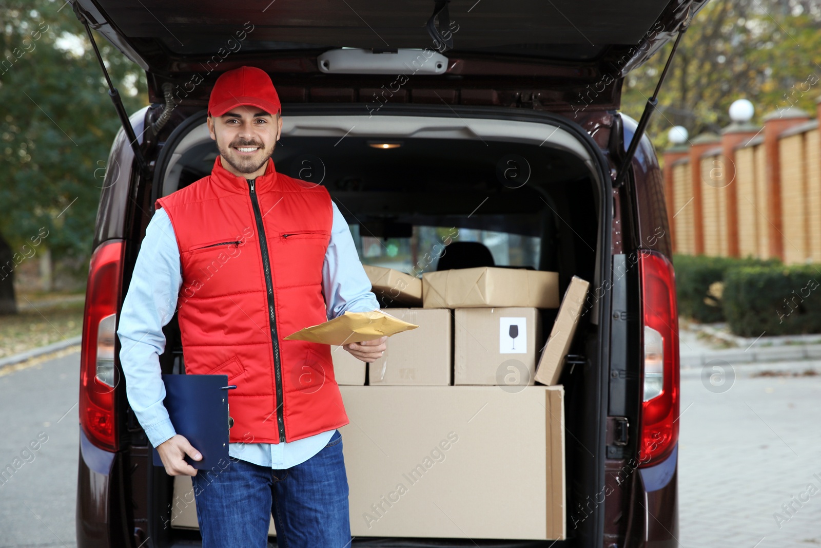 Photo of Young courier with parcel and clipboard near delivery van outdoors