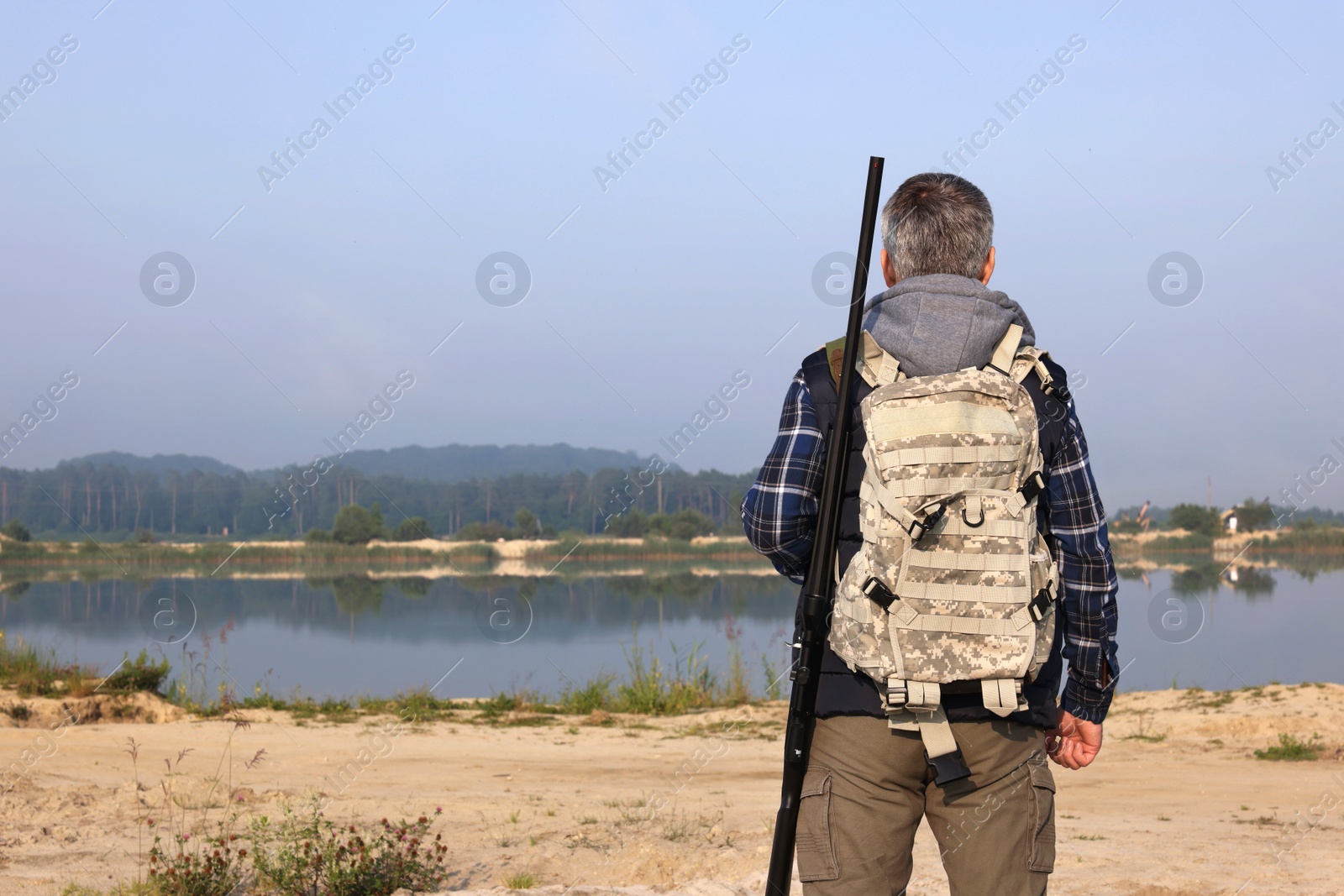 Photo of Man with hunting rifle and backpack near lake outdoors, back view. Space for text