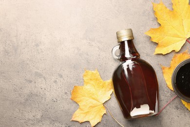 Photo of Bottle of tasty maple syrup, bowl and dry leaves on light grey table, flat lay. Space for text