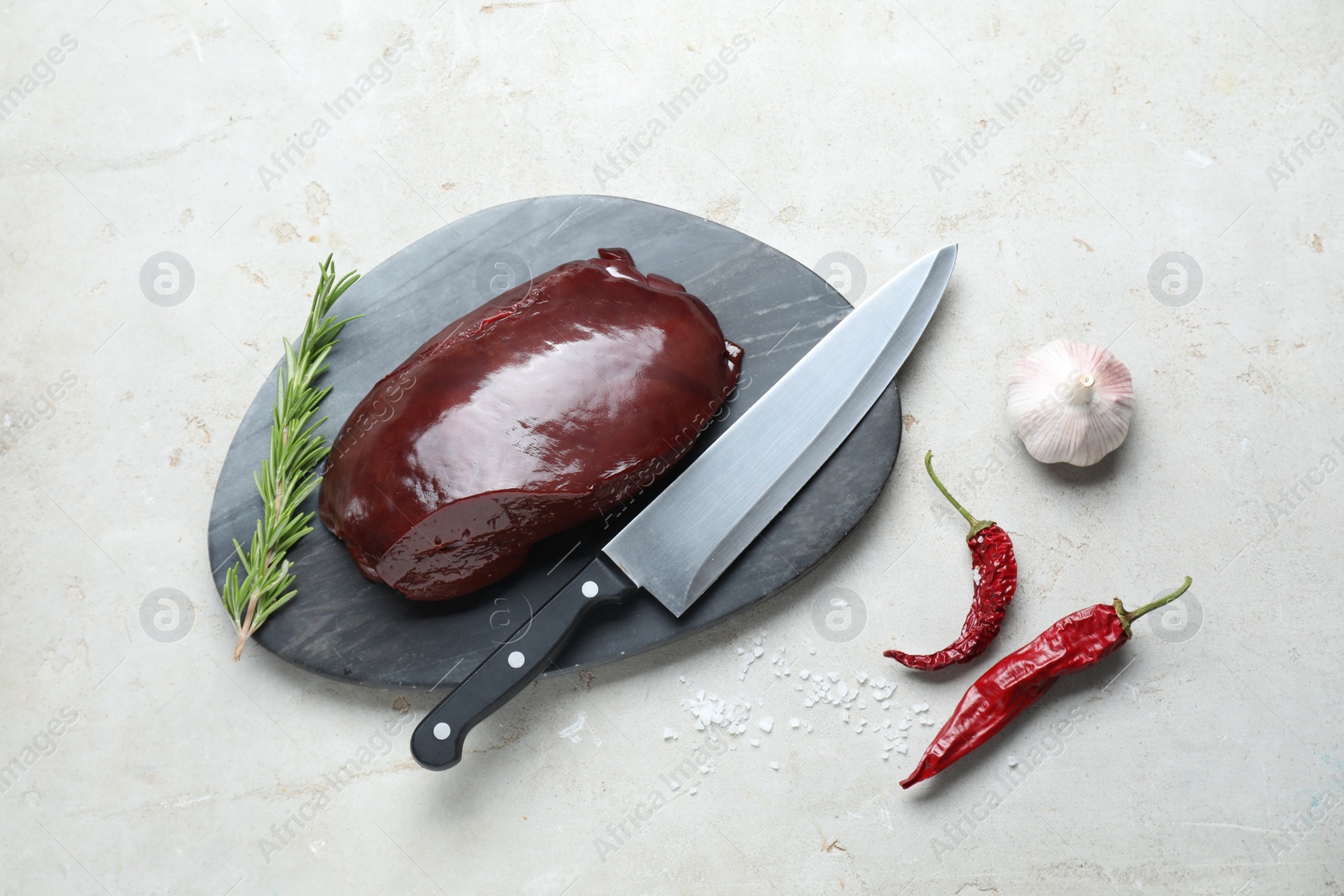 Photo of Piece of raw beef liver with chili peppers, rosemary, garlic and knife on light table, flat lay