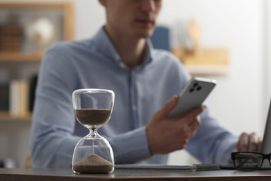 Photo of Hourglass with flowing sand on desk. Man using smartphone indoors, selective focus