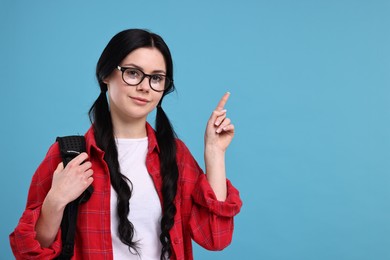 Photo of Student in glasses with backpack pointing at something on light blue background. Space for text