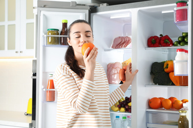 Young woman with orange near open refrigerator indoors