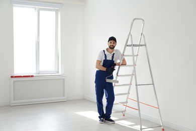 Full length portrait of handyman with roller brush near ladder indoors, space for text. Professional construction tools