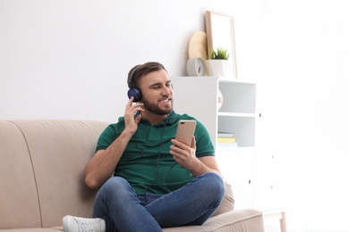 Photo of Young man in headphones with mobile device enjoying music on sofa at home