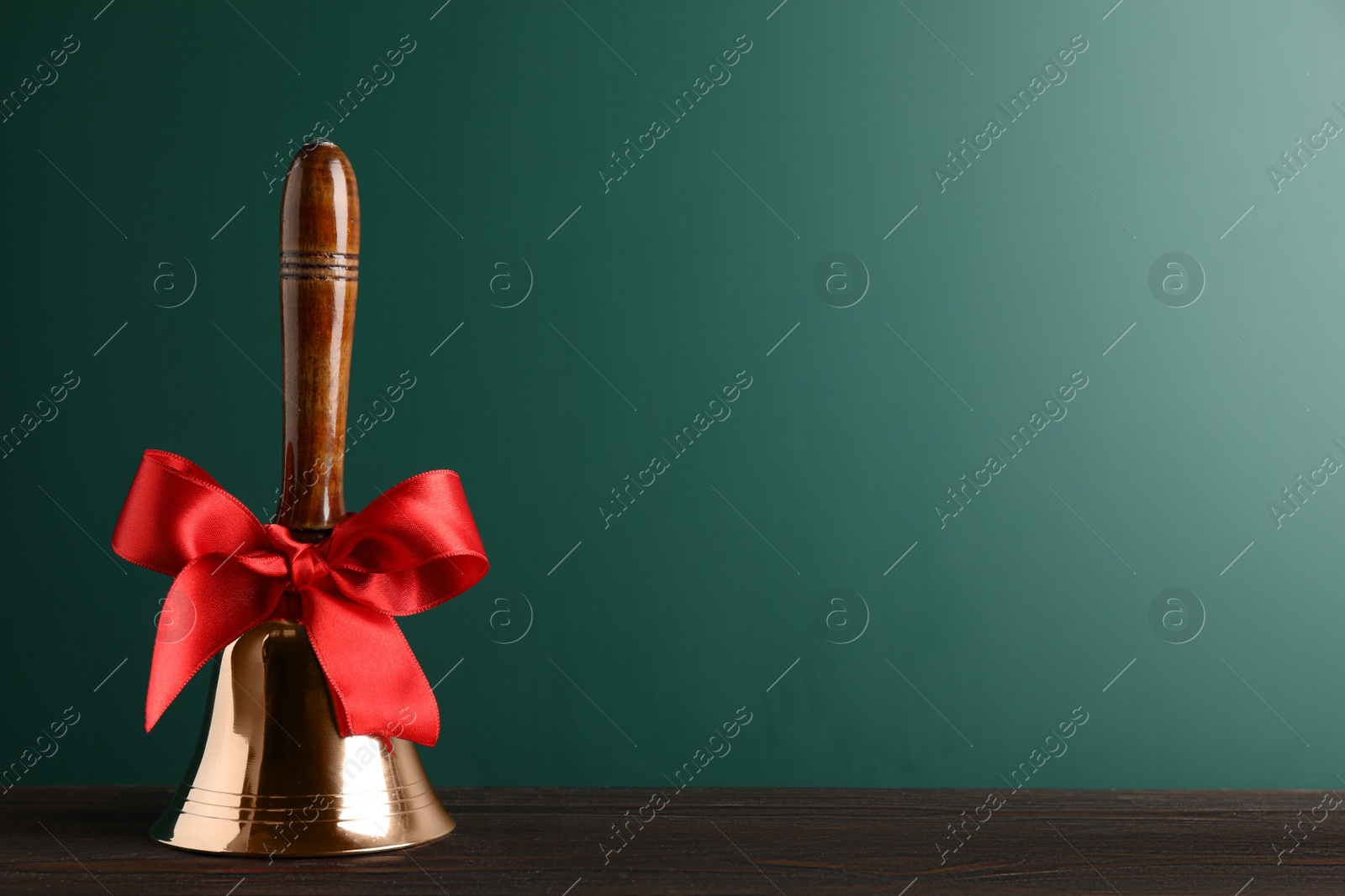 Photo of Golden bell with red bow on wooden table near green chalkboard, space for text. School days